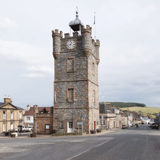 Uhrturm aus Bruchstein auf der Kreuzung einer schottischen Kleinstadt bei grauem Himmel