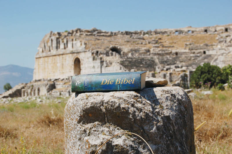 Eine blaue Bibel liegt auf einem Stein. Im Hintergrund die Theaterruine im antiken Milet in der Türkei.