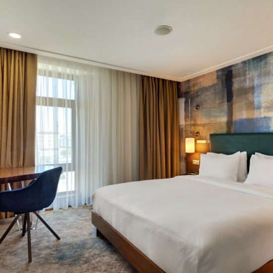 Hotelzimmer mit hellbrauenen Gardinen, weiß bezogenem Doppelbett, Sessel und einer Tapete in brauner Wischtechnik.