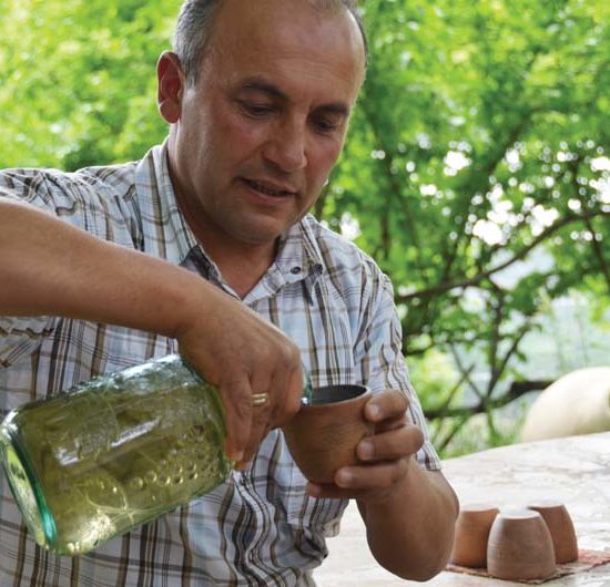 Ein Mann in einem hellen karierten Hemd gießt aus einer Flasche hellen Wein in einen Tonbecher.