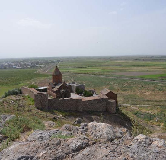 Eine steinerne armenische Klosteranlage mit Rundturm liegt hinter Festungsmauern an einem Hand. Dahinter liegen flache Felder.
