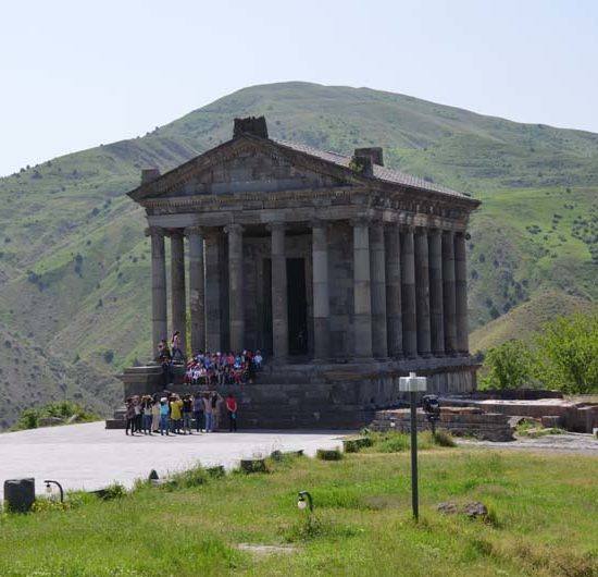 Im römischen Stil mit Säulen und einem Dreiecksgiebel erbauter Tempel Garni vor grünem Bergland und mit einer Gruppe Touristen vor dem Bauwerk.