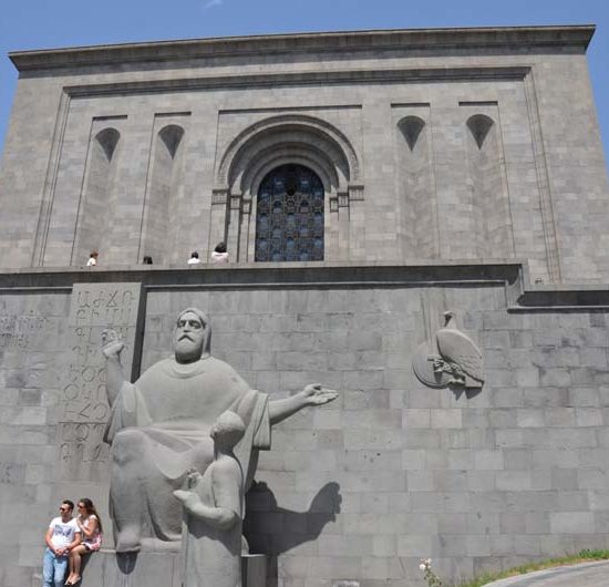 Monumentaler frauer Steinbau des Matenadaran Museums in Jerewan mit Statue, vor der ein Paar sitzt.