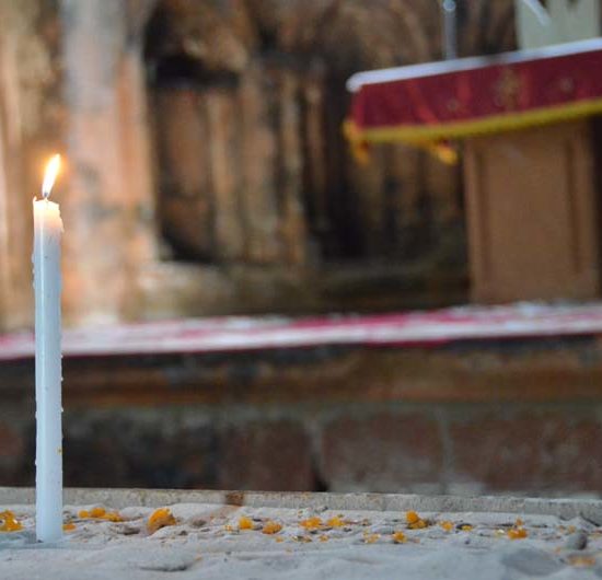 Im Kloster Marmaschen steckt eine weiße, schlanke, brennende Kerze im Sand. Im Hintergrund der Altar mit rotem Tuch.