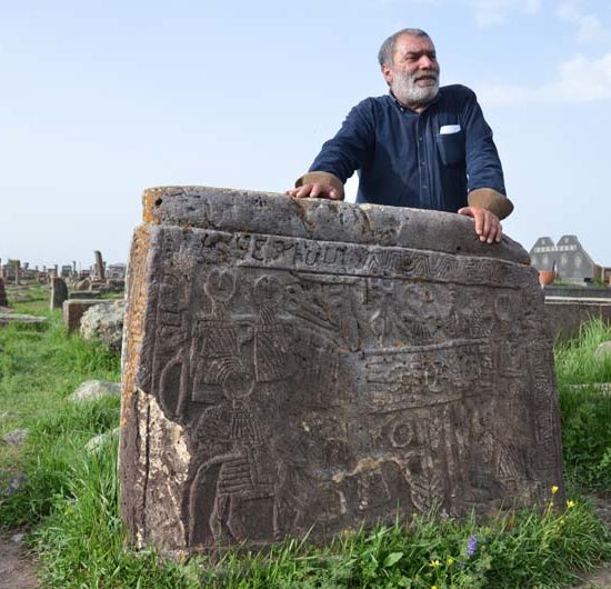 Ein Mann mit blauem Hemd steht auf dem Friedhof Noradus hinter einem Kreuzstein mit filigranem Relief. Im Hintergrund eine grüne Wiesen mit vielen Grabmalen.