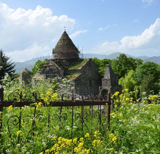 Die armenische Klosterkirche Sanahin inmitten von einer grünen Wiese und Wald, die die Kirche zu überwuchern scheinen. Im Vordergrund ein Eisenzaun mit Ornamenten und im Hintergrund die Berge Armeniens.