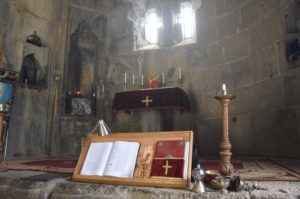 Chor des Tatev-Klosters, dessen Altar von einem roten Tuch mit goldenem Kreuz bedeckt ist. Im Vordergrund ein Halter mit aufgeschlagenem Buch und ein Kerzenständer.