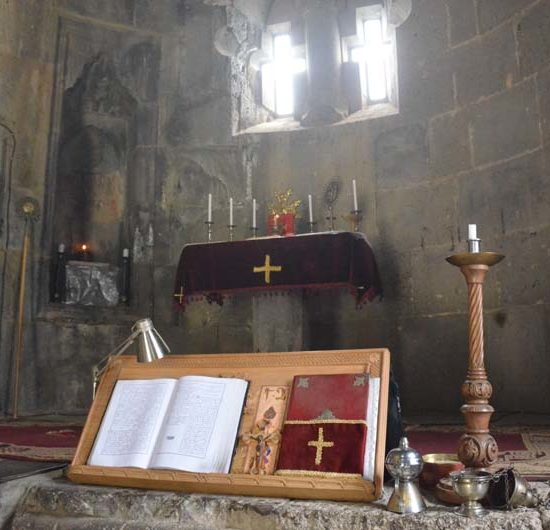 Chor des Tatev-Klosters, dessen Altar von einem roten Tuch mit goldenem Kreuz bedeckt ist. Im Vordergrund ein Halter mit aufgeschlagenem Buch und ein Kerzenständer.