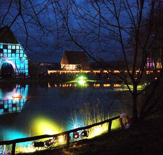 Nachtaufnahme mit farbiger Beleuchtung von an einem Teich gelegenen Fachwerkhäusern des Freilichtmuseums Detmold.