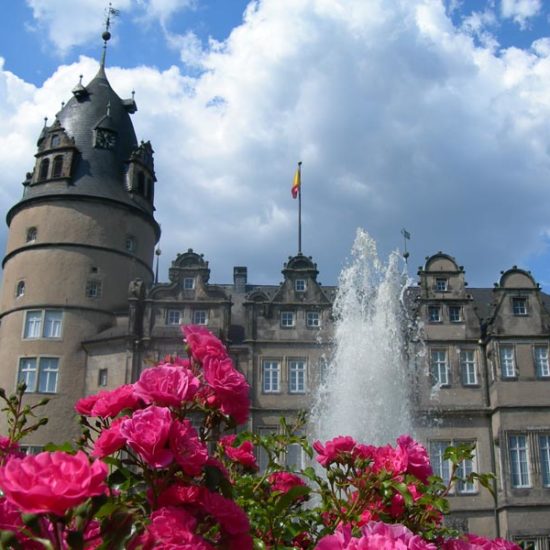 Renaissanceschloss Detmold mit Wehrturm und Elementen der Weserrenaissance. Im Vordergrund rosa Rosen und die Fontäne eines Springbrunnens.
