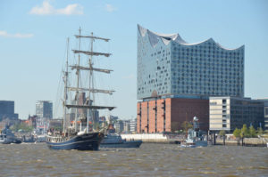 Backsteinkubus der Elbphilharmonie Hamburg mit modernem himmelblauem Aufbau direkt am Fluss Elbe, auf dem das Dreimasterschiff Thalassa fährt.- Foto © REISEKULTOUREN