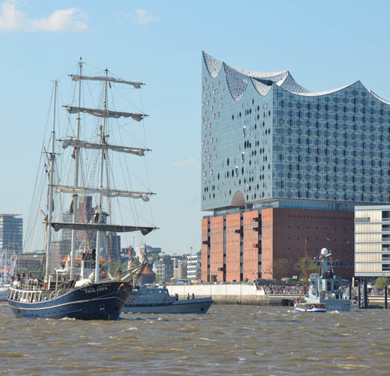 Backsteinkubus der Elbphilharmonie Hamburg mit modernem himmelblauem Aufbau direkt am Fluss Elbe, auf dem das Dreimasterschiff Thalassa fährt.- Foto © REISEKULTOUREN