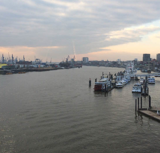 Abendstimmung mit Blick von oben auf den Hafen Hamburg und die Elbe. Rechts liegen weiße Schiffe an den Anlagern der Landungsbrücken, links Kräne der Werften und im Hintergrund drei Hochhäuser.