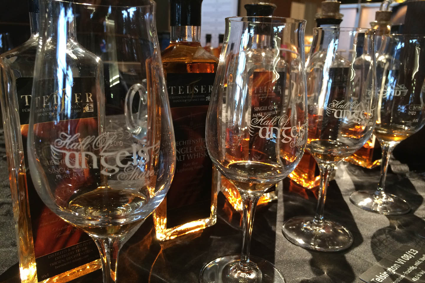 Vier Whiskygläser mit Stil und der Beschriftung Hall of Angels Share stehen vor einer Reihe von eckigen Whiskyflaschen der Marke Telser.