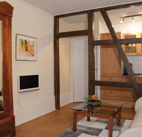 Wohnzimmer mit hellem Sofa, Tisch und antikem Schrank, durch eine offene Fachwerkwand abgegrenzt zu einer Küchenzeile mit Holzfronten.