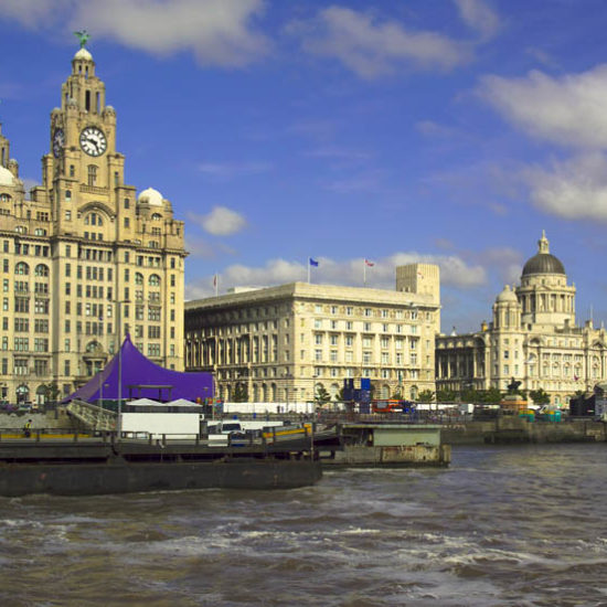 Drei als die "Three Graces" bezeichnete mehrstöckige, helle historische Steingebäude mit Türmchen und Uhr liegen an der Uferpromenade des River Mersey.