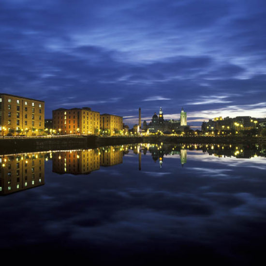 Tiefblaue Abendstimmung am Fluss rahmt gelblich beleuchtete Gebäude. Im Hintergrund die als Three Graces bezeichneten historischen Steinbauten mit Türmen.