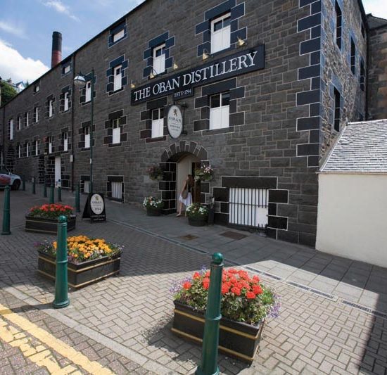 Dunkle Steinfassade der Oban Distillery mit Blumenkübeln im Vordergrund.