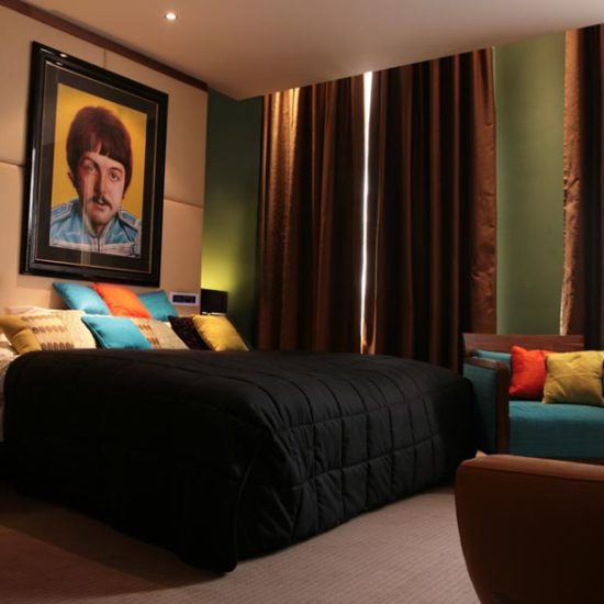 Hotelzimmer mit schwarzer Bettdecke, bunten Kissen, grünen Wände und brauner Gardiene. Über dem Bett hängt ein großes Portrait.