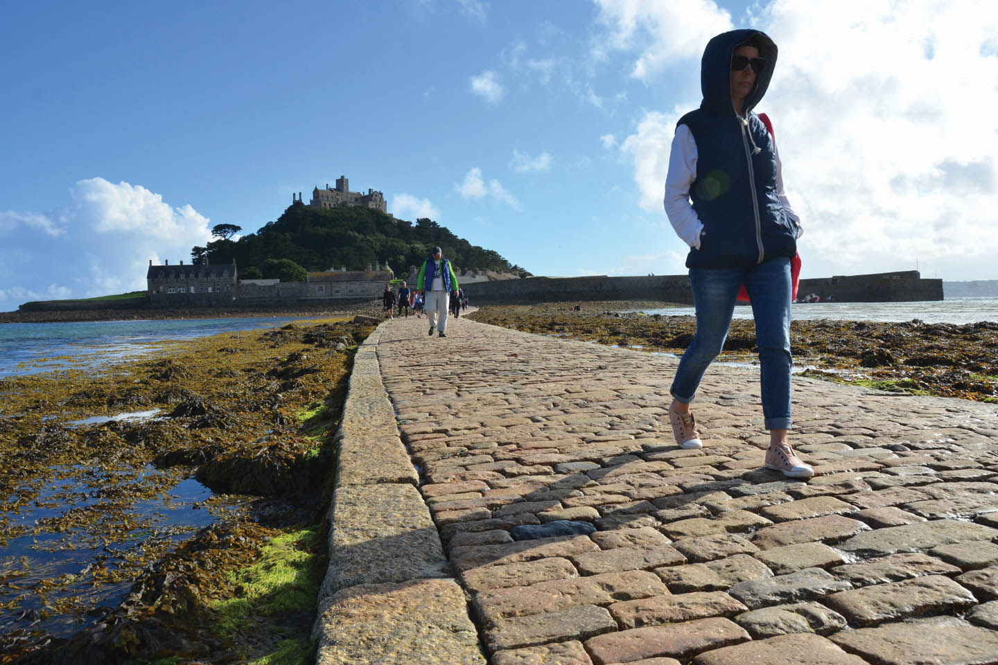 Menschen laufen bei blauem Himmel auf einem gepflasterten Causeway von der Burg St. Michael's Mount aus bei Ebbe durch das Meer.