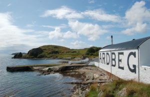 Weißes flaches Lagerhaus mit Ardbeg-Schriftzug liegt direkt an der felsigen Küste und dem Meer, in das ein Bootsanleger ragt.