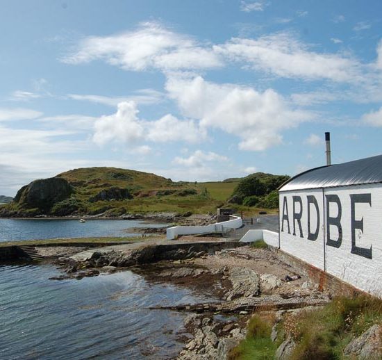 Weißes flaches Lagerhaus mit Ardbeg-Schriftzug liegt direkt an der felsigen Küste und dem Meer, in das ein Bootsanleger ragt.