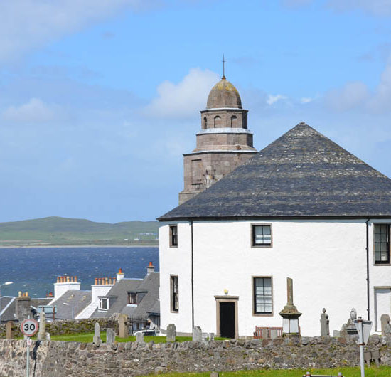 Die weiße Rundkirche von Bowmore mit rundem Glockenturm liegt inmitten eines Friedhofes oberhalb einer Meeresbucht.