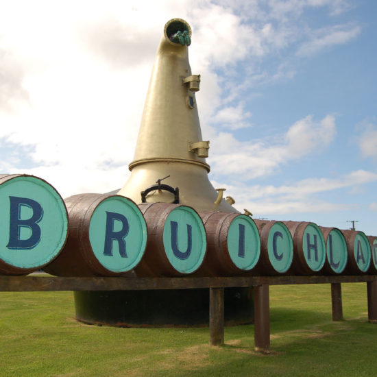 Schriftzug Bruichladdich, auf Fässer gepinselt, vor einer Kupferbrennblase in der oben Stiefel herausschauen.