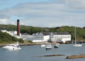 Die weißen Gebäude der Lagavulin Destillerie liegen in einer Meeresbucht, in der kleine Schiffe und Yachten ankern. Markant ist ein roter Turm mit schwarzer Spitze.