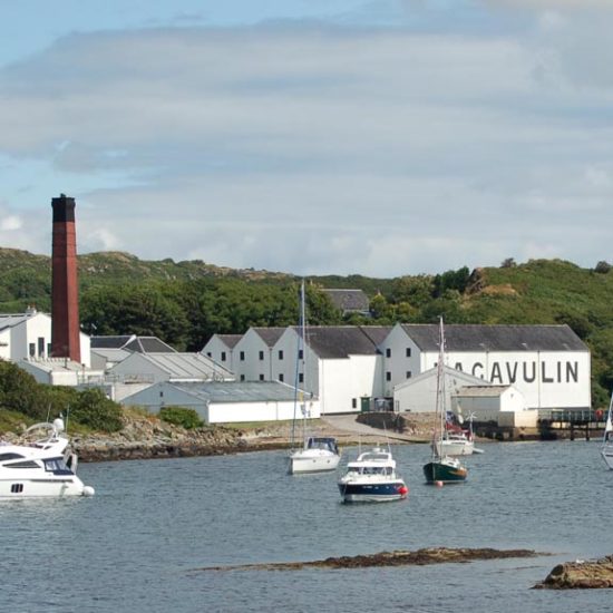 Die weißen Gebäude der Lagavulin Destillerie liegen in einer Meeresbucht, in der kleine Schiffe und Yachten ankern. Markant ist ein roter Turm mit schwarzer Spitze.