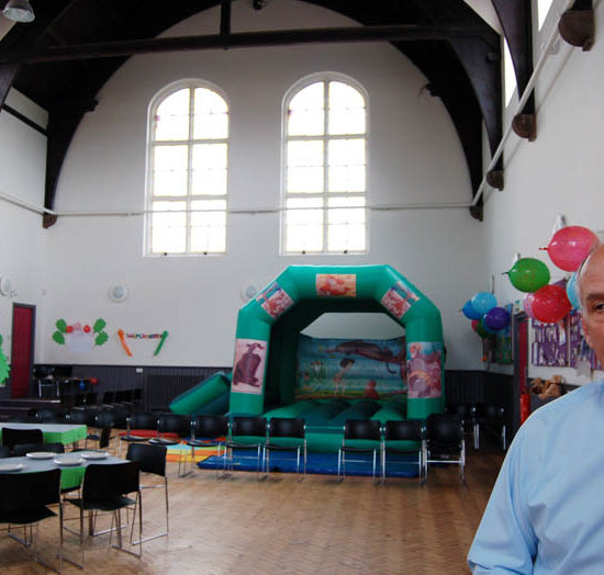 Mit Luftballons und Fotos geschmückter Gemeinderaum der St. Peter's Church Liverpool mit Tischen, Hüpfburg und einem Mann mit hellblauem Hemd.
