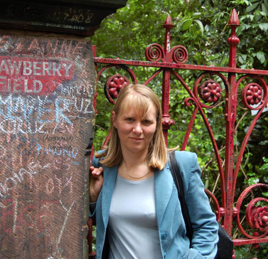 Blonde Frau mit türkisfarbenem Blazer lehnt sich vor einem erdbeerroten Gittertor an einen mit Strawberry Field beschriften Steinpfosten.