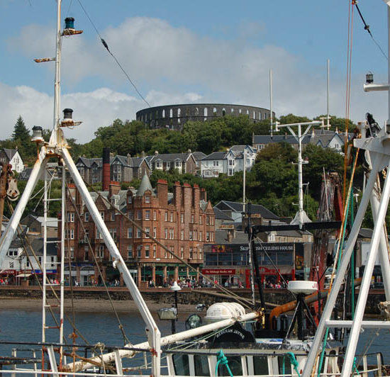 Blick über ein Schiff hinweg auf das Hafenbecken von Oban und ein rundes Fort auf dem Berg.