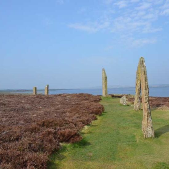 Monolithen stehen im Halbkreis als Felsreihe auf Gras und Heidefläche. Im Hintergrund blauer Himmel und Meer.