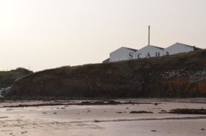 Die Lagenhäuser der Scapa Distillery überragen knapp die Klippen am Strand.