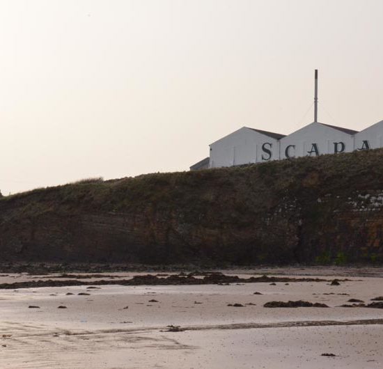 Die Lagenhäuser der Scapa Distillery überragen knapp die Klippen am Strand.