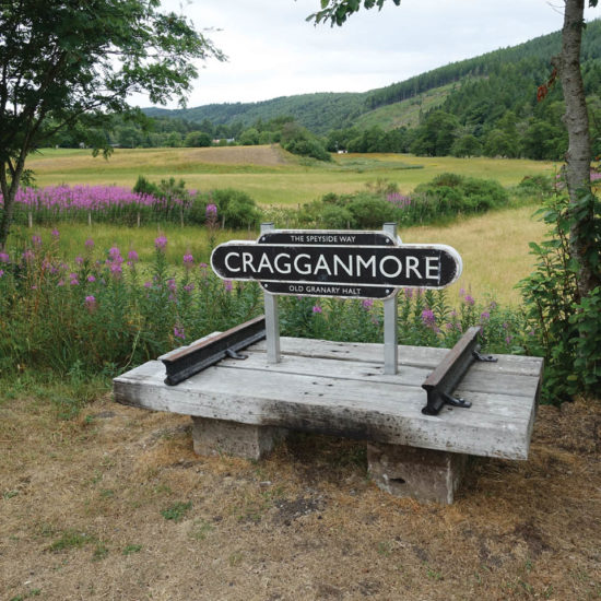 Craggammore-Schild auf einer Holzkonstruktion mit Bahnschienen. Im Hintergrund das grüne Tal des River Spey.