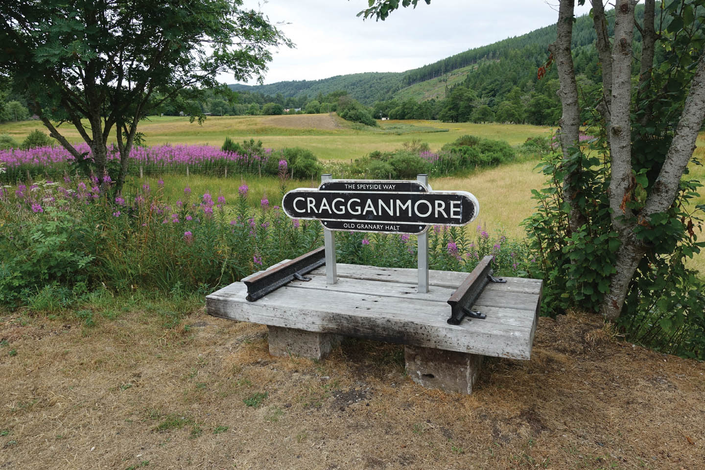 Craggammore-Schild auf einer Holzkonstruktion mit Bahnschienen. Im Hintergrund das grüne Tal des River Spey.