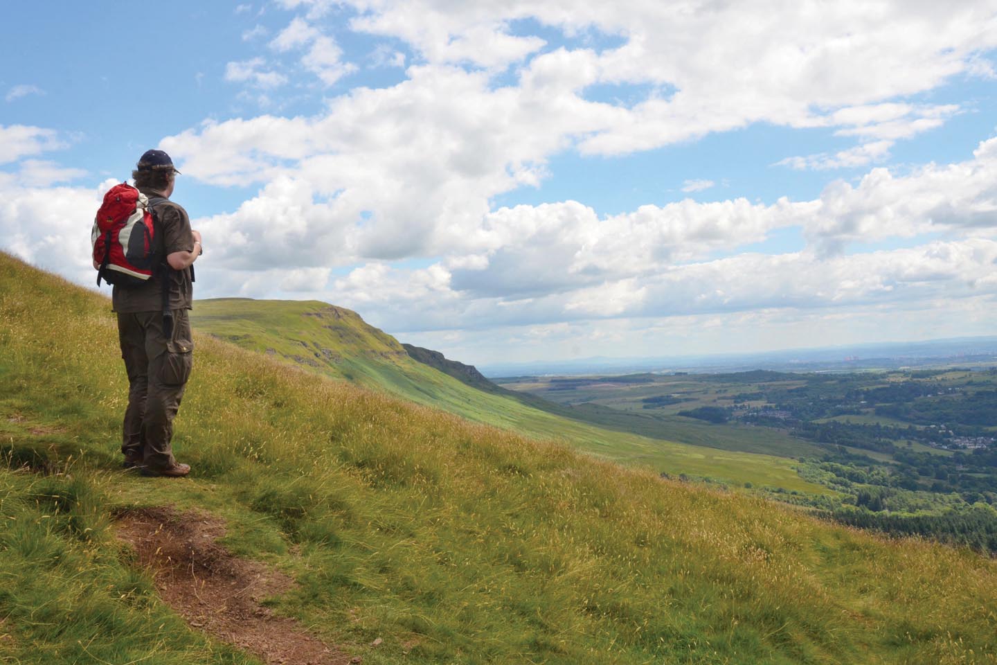 Wanderer mit Rucksack steht bei Sonnenschein an der Steilstufe der schottischen Highlands und schaut die grünen Hänge hinunter in die Lowlands.