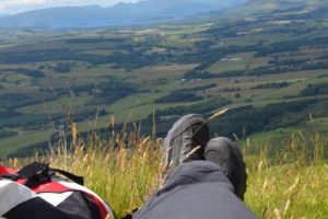 Blaue Wanderschuhe und roter Rucksack an einer Bergkante mit Blick tief hinunter in die grüne Ebene mit dem Loch Lomond und den Bergen der Highlands im Hintergrund.