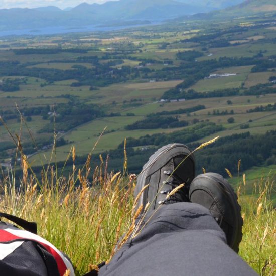 Blaue Wanderschuhe und roter Rucksack an einer Bergkante mit Blick tief hinunter in die grüne Ebene mit dem Loch Lomond und den Bergen der Highlands im Hintergrund.