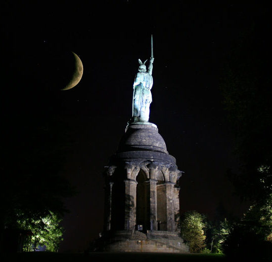 Nachtaufnahme mit dem angestrahlten Hermannsdenkmal, einer Kufterstatue mit Schwert auf hohem Sockel, vor einer gelben Mondsichel.