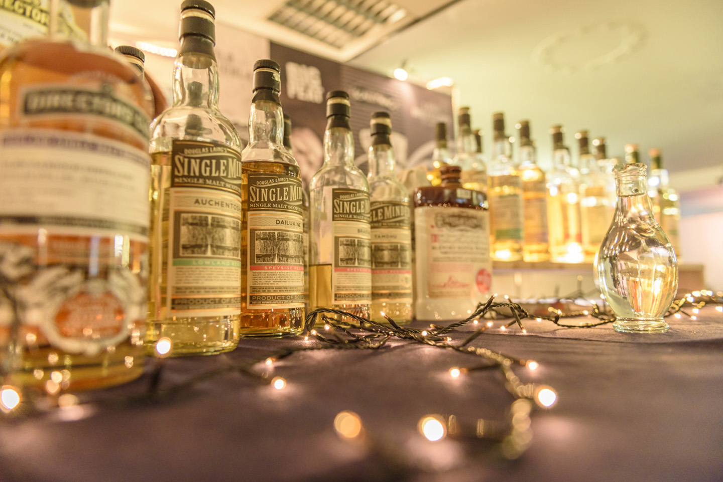 Eine Reihe fast gleicher Single Malt Whiskyfalschen steht auf einem Tisch mit Lichterkette und einem Glaskrug mit Wasser.