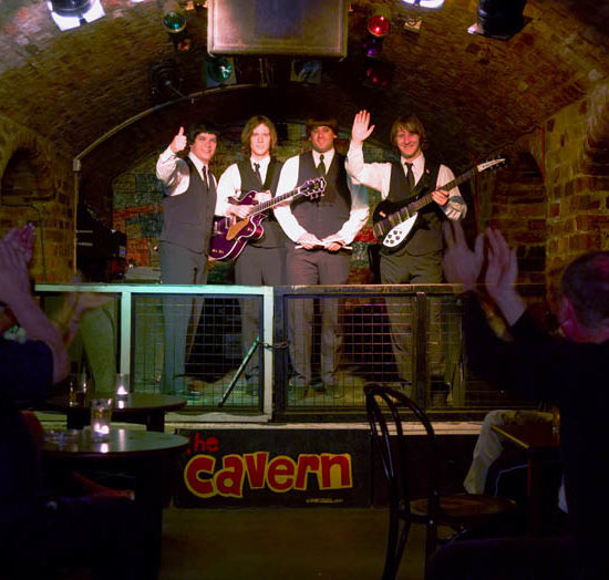 Bühne im Gewölbekeller des Cavern Club Liverpool. Darauf winken vier Musiker in dunklen Anzügen und mit weißem Hemd dem Publikum zu.