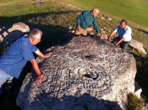 Drei Männer stehen auf einer Wiese um einen großen flachen Felsen herum und diskutieren dessen Relief, das als Sternenkarte gedeutet wird.