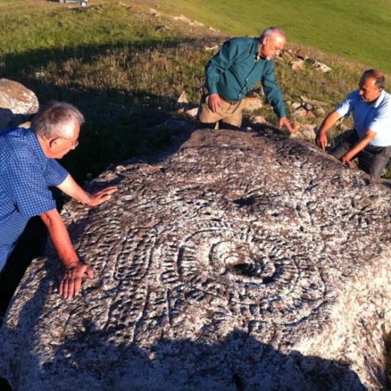 Drei Männer stehen auf einer Wiese um einen großen flachen Felsen herum und diskutieren dessen Relief, das als Sternenkarte gedeutet wird.