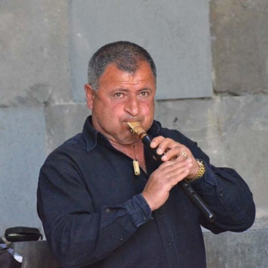 Mann mit kurzen Haaren und blauem Hemd spielt vor einer Steinmauer auf einer armenischen Duduk Flöte.