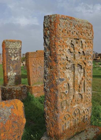 Vier mit gelbbraunen Flechten und reichen Steinreliefs überzogene Kreuzsteine stehen hintereinander auf einem Friedhof und glänzen vor blauem Himmel im Sonnenlicht.