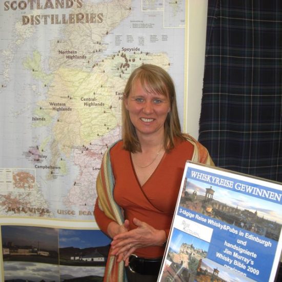 Frau mit halblangen blonden Haaren und orange Shirt vor einer Whisky-Landkarte Schottlands.
