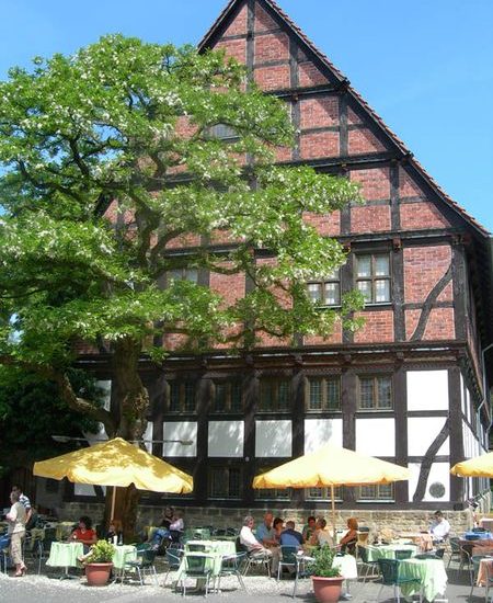 Stattliches Fachwerkhaus hinter großem Baum und Außengastronomie mit gelben Sonennschirmen.